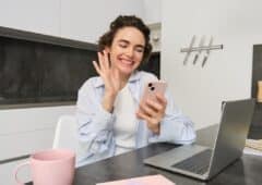 Happy young woman says hello, waves hand at smartphone and smiles, video chats with someone on mobile phone app